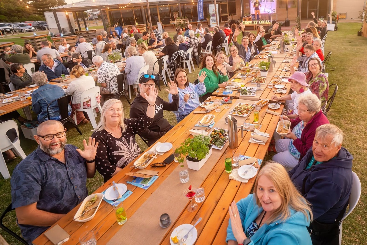 A group of people seated at an outdoor dining table, all looking at the camera smiling and waving.